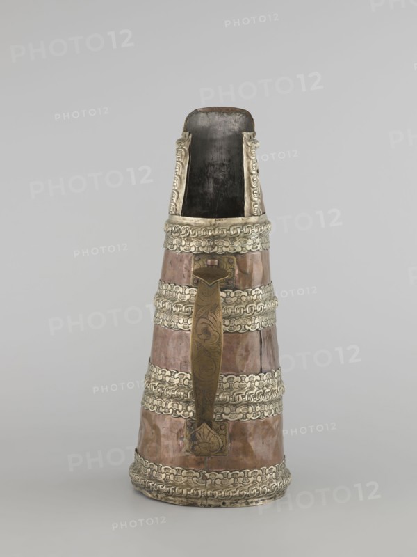 Tea pot, 19th century, Tibet. Teapot/kettle; vessel tapering towards the top with brass handle and silver fittings. Height 35.0 cm; width 19.0 cm; diameter 15.0 cm. Tibetan craft, tea vessel., An upright conical vessel with a narrow open top and a wider base, featuring a metallic brown body accented by several horizontal decorative bands of silvery metal; a long vertical handle of darker brass-like color with engraved patterns runs down the front and is attached near the top and bottom; the interior visible at the top is dark, and the silvery bands and rim display embossed interlocking designs; the overall colors visible are brown, silvery gray, dark brass, and dark gray.