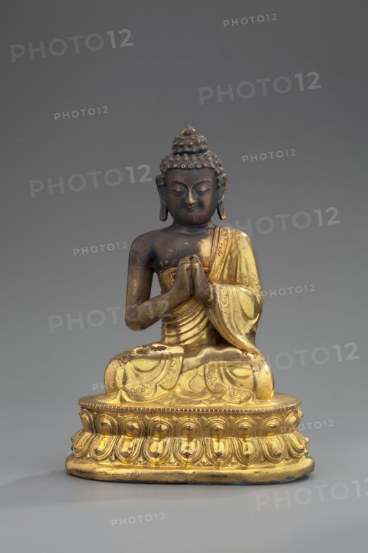 Shakyamuni Buddha, unknown sculptor, Himalayan, Sino-Tibetan style, Tibetan Buddhist art; Buddha ca. 560–480 BCE, ushnisha, lotus throne, mudras; 18th century, Tibet; sculpture, figurine; height 11 cm, width 7.7 cm., A seated statuette of a robed figure with hands held together in front, rendered in a combination of dark brown and gold tones; the figure has a textured hair bun and elongated earlobes, a serene facial expression, and detailed drapery on the robe; the base is ornate with carved petal-like patterns and is predominantly gold with some worn darker spots; the background is a smooth gray.