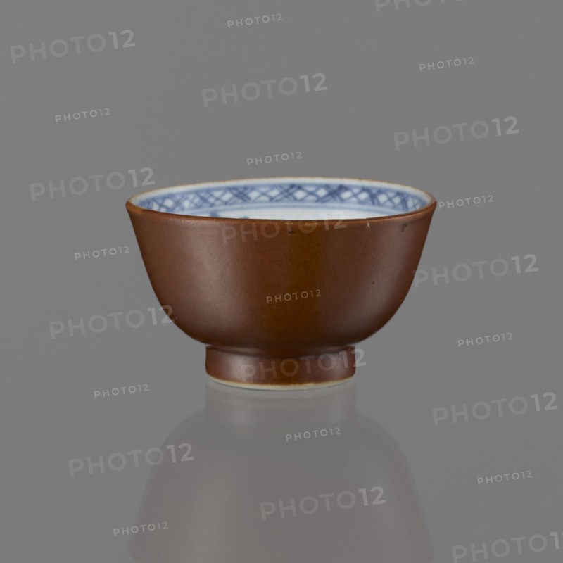 Alcohol bowl, Chinese art, Chinese culture, Qing 1644–1911, ceramics, blue-and-white porcelain, landscape motifs, 19th century, China, vessel, height 4.5 cm, diameter 7.8 cm, A small ceramic bowl with a smooth brown exterior and a glossy finish sits centered on a gray surface, showing a few subtle imperfections on its outer wall; the interior rim displays a decorative pattern in blue on a white background, and a narrow lighter band appears at the base where the bowl meets the surface, with a soft reflection of the bowl visible below.