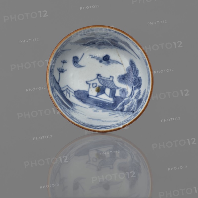 Alcohol bowl, Chinese, Qing dynasty 1644–1911, 19th century. Blue-and-white porcelain vessel with landscape motifs; height 4.5 cm, diameter 7.8 cm., A shallow circular ceramic dish with a brown rim sits against a gray background, showing a blue painted scene inside that includes a small building with a pitched roof, surrounding trees, a few clouds, and stylized landscape brushwork; the dish casts a soft, muted reflection below it that mirrors the blue-and-white interior design and brown edge.