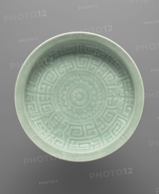 Plate - celadon, Chinese, Chinese art, lotuses, dragons iconography, ceramics, 18th century, China, vessel, height 5.4 cm, diameter 28.5 cm, A shallow round ceramic dish shown from above with a pale celadon green glaze featuring a raised central floral rosette surrounded by a geometric maze-like band and decorative corner motifs, with the rim and interior displaying subtle variations of light green and mint tones against a neutral gray background.