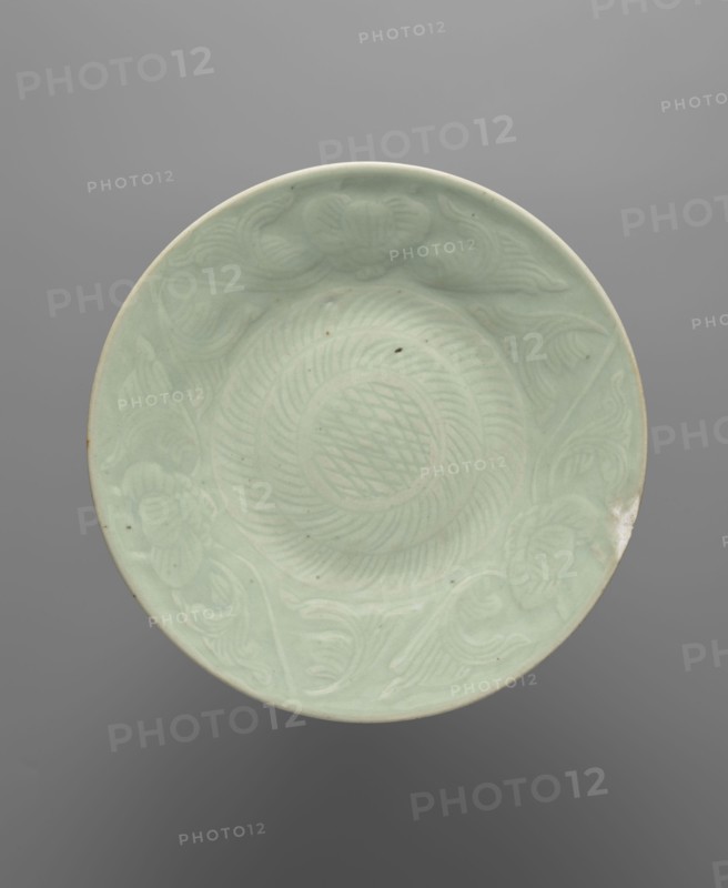 Plate — celadon; maker, Chinese; ceramics; geometric decorations and plant ornaments; vessel; 13th–15th century, China; height 3.5 cm; diameter 27.2 cm., A round shallow dish with a smooth pale green surface and a slightly raised rim, featuring carved concentric circular patterns and flowing carved motifs around the inner edge; the glaze shows tiny dark specks and a small area of worn or discolored edge, all set against a neutral gray background.