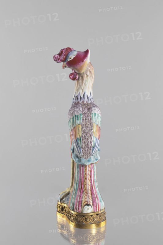 Bird, Chinese, Qing 1644–1911, 18th/19th century. Porcelain figure of a bird pheasant/phoenix, fenghuang iconography in colored porcelain on a brown gilded openwork base with braided ornament and rosettes. Sculpture; height 40.4 cm, width 10.3 cm., A ceramic bird sculpture standing upright on an ornate golden base, with a head turned to the side and a curved beak; the head and crest show shades of deep pink, magenta, and small areas of pale yellow, while the neck has cream and light beige tones blending into soft lavender. The body features a textured pattern of feathers in muted purple, teal, and turquoise with accents of pale yellow and coral, and the wings exhibit a mix of blue and green hues. The tail and lower body display vertical fluted stripes in pink, teal, and cream with subtle gold detailing, and the gilded base has intricate carved patterns in a warm metallic gold. The background is a uniform light gray.