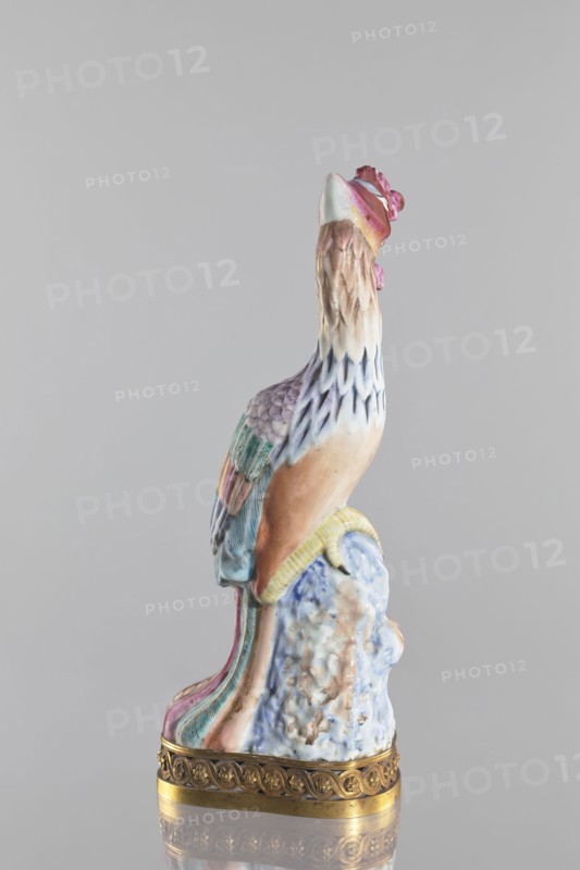 Bird, Chinese, Qing dynasty 1644–1911; porcelain figure of a bird pheasant/phoenix, fenghuang iconography in colored porcelain on a brown gilded openwork base with braided ornament and rosettes; 18th–19th century, China; sculpture; height 40.4 cm, width 10.3 cm., A ceramic sculpture of a bird perched on a rocky base, shown in profile, with a crest and elongated tail; the bird displays pastel colors including pink, blue, green, beige, brown, and white, with layered textured feathers on the neck and body and multicolored tail feathers that flow down the base; the rocky base shows mottled blue, white, and brown tones and sits on a decorative metallic rim in gold.