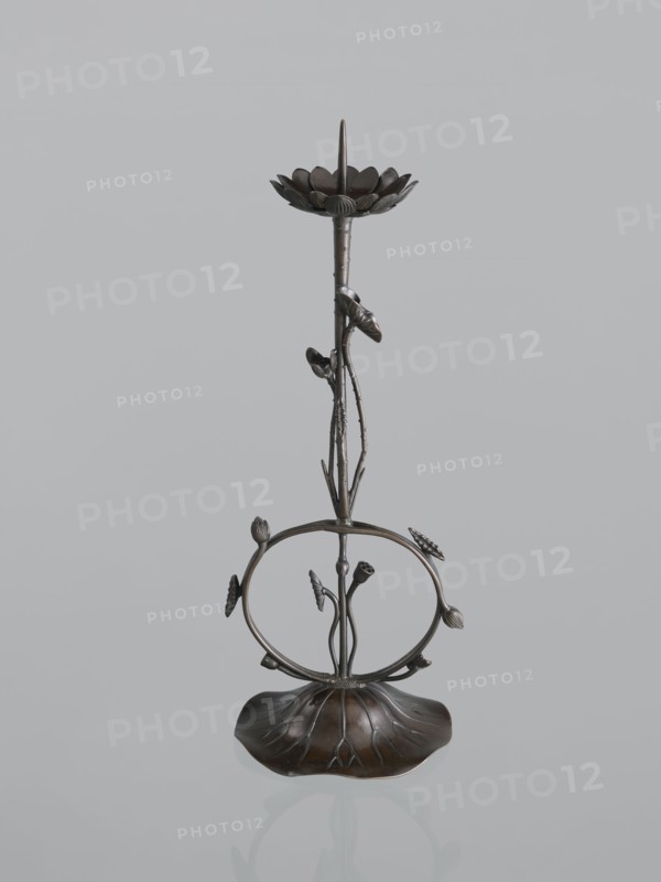 Candlestick shokudai, maker: Takimoto Iwami, Japanese; motifs: lotuses and leaves; Meiji period 1868–1912, 2nd half of the 19th century; Japan; medium: candlestick; height: 46.0 cm; provenance: unknown., A slender metallic sculpture with a dark brown and bronze finish stands against a pale gray background; the piece features a petal-like dish at the top with a pointed element rising from its center, a thin textured stem with several small bud-like forms attached, a circular open ring midway adorned with leaf and bud shapes, and a broad, curved base shaped like a stylized leaf that reflects slightly on the surface beneath.