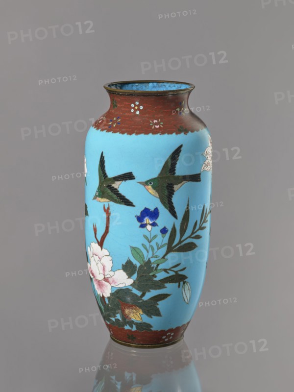 Vase, Cloisonné enamel on copper with brass rims at the spout and foot. Short, slightly flared neck. Decoration on a blue background: large motifs of rose bushes and lilies, small flowers, two butterflies and a bird kachôga. Japanese, Meiji period, ca. 1905–1908. Vessel; height 30.7 cm, diameter 10.6 cm., A tall cylindrical vase with a rounded shoulder and flared rim sits against a neutral gray background; the main body is a bright sky blue with painted motifs including a group of green and black birds in flight and floral arrangements featuring white and pink blossoms, a blue flower, and dark green leaves, with some reddish-brown branches. The neck and base areas have a band of mottled reddish-brown decorated with small floral accents, and the interior rim shows a darker blue. The vase casts a soft reflection on the surface below.