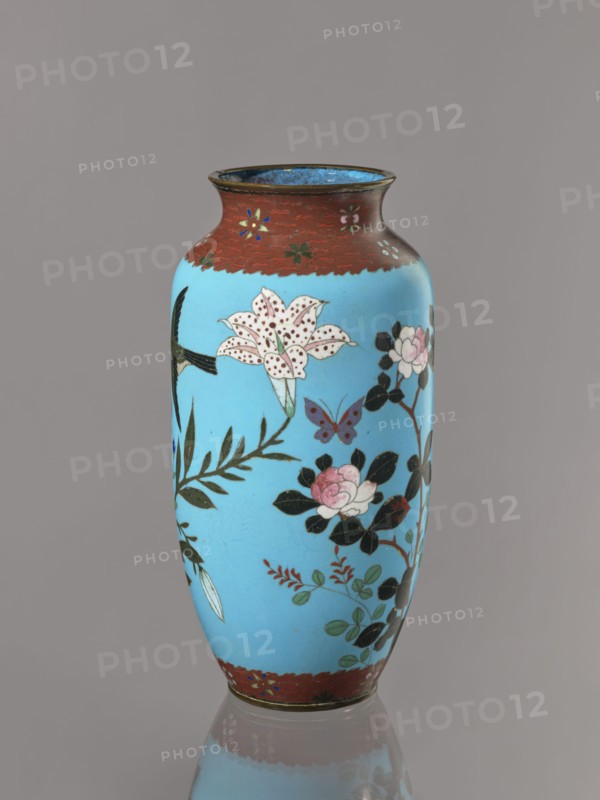 Vase, Cloisonné enamel on copper with brass rims at the spout and foot. Short, slightly flared neck. Decoration on a blue background: large motifs of rose bushes and lilies, small flowers, two butterflies and a bird. Iconography: flowers, butterflies, roses, lilies, birds kachôga. Japanese, Meiji period, ca. 1905–1908. Vessel; height 30.7 cm, diameter 10.6 cm., A tall vase with a rounded body and slightly flared rim sits against a plain background; the vase is primarily bright blue with decorative bands of reddish-brown at the top and bottom, and features painted floral motifs including a large spotted white lily, pink rose-like flowers, dark green leaves, a small purple butterfly, and other leafy sprigs in shades of green and red, with areas of black used for stems and leaf accents and a thin metallic rim at the mouth.