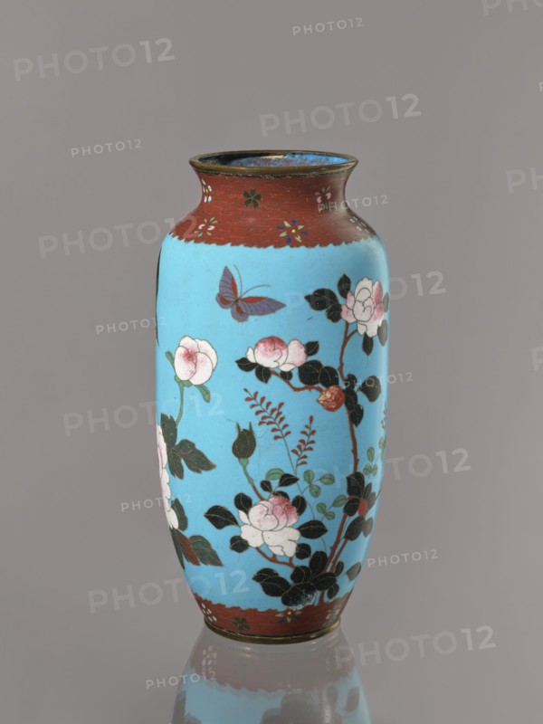 Vase. Cloisonné enamel on copper with brass rims at the spout and foot. Short, slightly flared neck. Decoration: large roses and lilies, small flowers, two birds and a butterfly on a blue background kachôga. Japanese, Meiji period, ca. 1905–1908. Height 30.8 cm; diameter 10.6 cm., A tall vase with a bright sky blue body and dark reddish-brown rim and base, decorated with painted floral branches bearing pale pink and white blossoms and dark green leaves, a few buds, and a single purple and red butterfly near the upper area, all set against a neutral gray background with a subtle reflection of the vase at the bottom.
