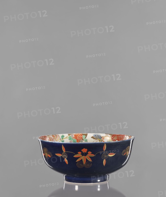 Bowl, Japanese, Arita, 2nd half of the 19th century. Colored porcelain vessel decorated with lotuses, fantasy animals, butterflies, flowers, and peonies; fo iconography. Height 10.8 cm, diameter 24.5 cm., A dark blue ceramic bowl with a glossy finish sits on a reflective surface. The bowl has an outward-flaring rim and a short foot. The exterior shows a pattern of stylized floral motifs in orange and gold with thin gold outlines and spiral accents. The interior rim reveals a multicolored decorative band with visible greens, reds, blues, and gold against a light background. The overall visible colors include dark blue, orange, gold, green, red, blue, and a light cream.