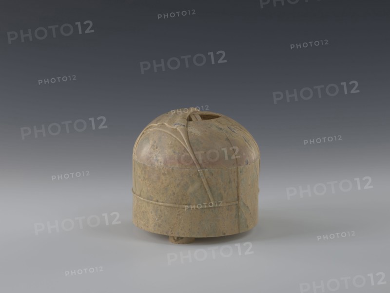 Tin with lid; maker, gift; Qing dynasty 1644–1911; 19th century; China; yurt decoration; container; height 12.8 cm; diameter 13.1 cm., A small rounded object sits on a smooth surface against a gradient background, appearing dome-like with a flat lower band and a slightly raised base; its surface shows a mottled mix of tan, beige, brown, and hints of green with areas of darker and lighter mottling, and there are carved or raised linear ridges and curved seams on the dome that create segmented panels and a small opening at the top.