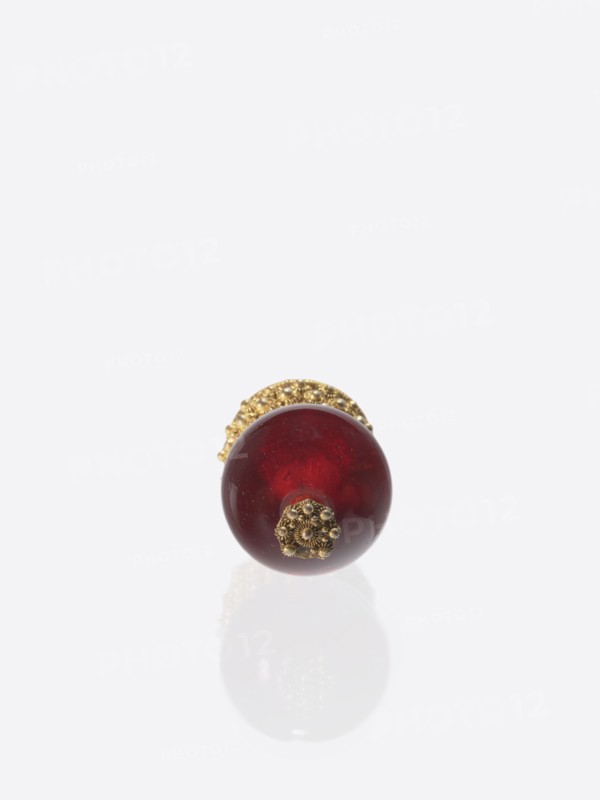 A button for a semi-official cap of a first-ranking official, Chinese, Qing dynasty 1644–1911. A button composed of a glass transparent ruby glass ball mounted on a pin with a dome decorated with rosettes. 19th century, China. Function: clothing/jewelry. Height 6.6 cm; diameter 3.0 cm., A single spherical object with a deep red glossy surface sits centered near the lower area of the image, topped with ornate metallic decorative elements in a gold color; the object casts a soft pale reflection on the light gray background, and small highlights and subtle surface imperfections are visible on the red sphere.