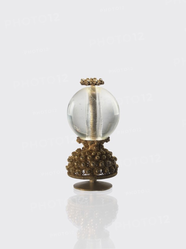 Button for a semi-official cap of a fifth-ranking official, Chinese, Qing dynasty 1644–1911. Button composed of a transparent glass ball mounted on a pin with a dome decorated with rosettes. 19th century. Materials: glass and metal. Function: cap button for an official. Dimensions: height 6.2 cm; diameter 2.9 cm., An ornate object sits against a pale gray background, consisting of a clear spherical glass orb perched on a decorative metallic base; the glass is transparent with subtle internal reflections and a central vertical element visible inside, and the metal base is a warm golden-bronze color with an intricate clustered beaded texture and floral-like motifs, topped by a small ring of matching beaded metal and standing on a round flat bronze pedestal that casts a soft mirrored reflection below.
