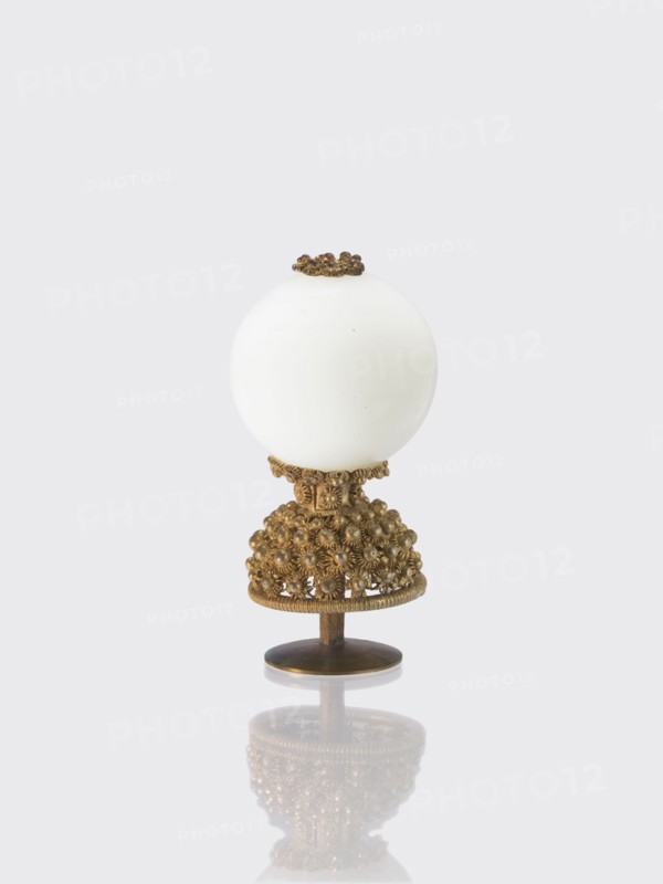Button for a semi-official cap of a sixth-ranking official., Chinese art; Chinese culture. Qing dynasty 1644–1911. Officials; emblems. Button composed of a milky-white glass ball mounted on a pin with a dome decorated with rosettes. 19th century, China. Clothing, jewelry. Height 6.4 cm; diameter 3.2 cm., A decorative object composed of a smooth white spherical top mounted on an ornate metallic stand, with the stand featuring an intricate cluster of small rounded decorative elements and filigree details in a warm golden-bronze color, a small circular base in a darker bronze supporting the piece, and a subtle reflection of the object visible on the glossy surface beneath; visible colors include white, golden-bronze, darker bronze, and pale gray in the background.