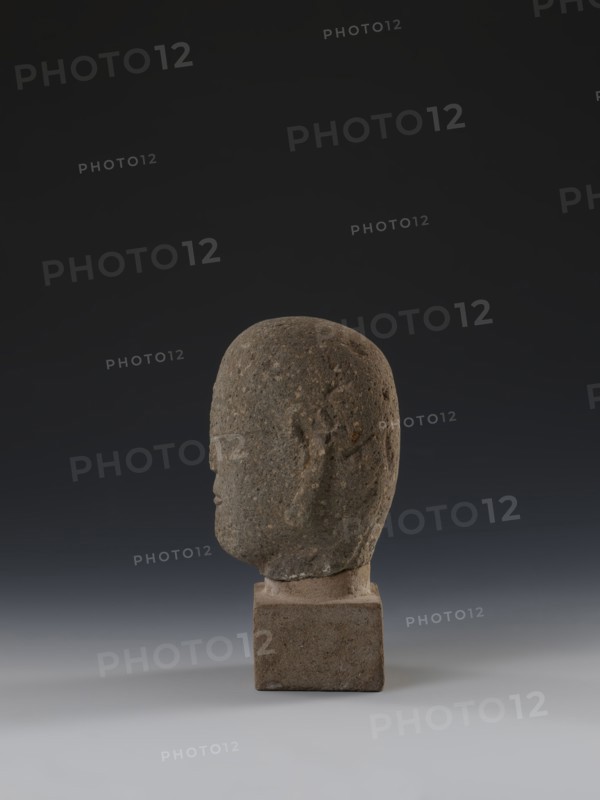 Head of a monk Buddha?, buddha iconography, Chinese art, Chinese culture, Ming 1368–1644, Chinese Buddhism, arhat iconography, Luohan iconography, 14th–15th century, China, sculpture, height 11.8 cm, width 9.2 cm, depth 8.7 cm, A weathered stone head sculpture in profile sits on a square stone base against a dark gradient background. The sculpture is textured with a rough surface showing small pits and irregularities, and the overall color palette includes shades of gray, brown, and muted beige on the stone, with the base a slightly warmer brownish-gray. The background transitions from nearly black at the top to a softer gray near the bottom, and the surface beneath the base is a light gray. The head has subtle carved facial contours visible from the side, including a rounded cranium, a brow line, a nose, and a hint of lips, with shadowing that gives a sense of depth.