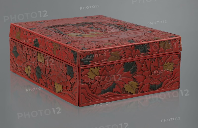 Box with a lid, Chinese art, Chinese culture, figural scenes, floral motifs, Qing 1644–1911, peonies, bamboos, geometric ornament, gourds, sages, scholars, fans, men. Decoration: on the lid a large reserve with a figural scene in a landscape; the rest of the background and sides decorated with floral motifs. Red, black, green, and yellow lacquer, carved. 19th century, China. Container. Height 13.0 cm, width 36.5 cm, depth 28.5 cm., A rectangular carved box sits on a reflective surface, shown from a low angled view. The box is predominantly red with carved floral patterns covering its top and sides, and a few carved leaves and petals are colored dark green and muted yellow. The carved surfaces have layered, sculpted textures with overlapping flower motifs and curved veining. The lid fits over the base with a narrow rim visible, and the reflection of the box appears faintly on the smooth surface beneath. The background is a uniform cool gray.