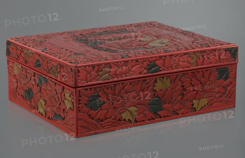 Box with a lid, Chinese art, Chinese culture, figural scenes, floral motifs, Qing 1644–1911, peonies, bamboos, geometric ornament, gourds, sages, scholars, fans, men. Decoration: on the lid a large reserve with a figural scene in a landscape; the rest of the background and sides with floral motifs. Red, black, green, and yellow lacquer, carved. 19th century, China. Container. Height 13.0 cm; width 36.5 cm; depth 28.5 cm., A rectangular decorative box sits on a reflective surface, covered in carved floral relief. The box is predominantly red with accents of dark green and muted yellow on the carved leaves and petals. The lid and sides show dense, overlapping flower and leaf motifs with textured, raised carving and smooth recessed background. The edges and corners of the box are finished in the same red, and the reflection below shows a softened mirror image of the box and its colors. The background behind the box is a flat neutral gray.