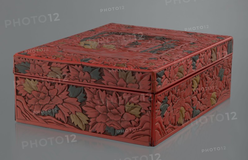 Box with a lid, Chinese art, Chinese culture, figural scenes, floral motifs, Qing 1644–1911, peonies, bamboos, geometric ornament, gourds, sages, scholars, fans, men. Decoration: on the lid a large reserve with a figural scene in a landscape; the rest of the background and sides with floral motifs. Red, black, green, and yellow lacquer, carved. 19th century, China. Container. Height 13.0 cm, width 36.5 cm, depth 28.5 cm., A rectangular carved box with a lid is shown against a neutral gray background and reflective surface; the box is covered in dense floral carvings with layered petals and leaves, with a dominant red color and accents of green and yellow on the carved foliage, and darker recessed areas that create shadowed texture across the top and sides.