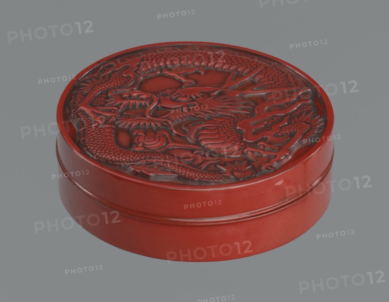 Box with a lid., Japanese, Meiji period 2nd half of the 19th century. Wooden core covered with lacquer; carved red lacquer exterior, black lacquer interior. Decoration: dragons, dragon with a pearl. Function: container. Dimensions: height 3.5 cm; diameter 10.9 cm; weight 149 g., A round lacquered box with a raised carved lid showing a coiled dragon motif, the box and carving predominantly red with darker red and black shading in the carved recesses; the exterior sides are smooth and glossy red; the background is a flat gray.