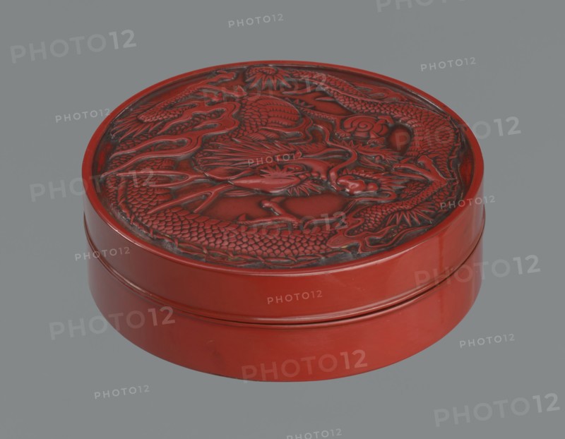 Box with a lid. Japanese, Meiji period 2nd half of the 19th century. Wooden core covered with lacquer; carved red lacquer on the outside, black lacquer on the inside. Decoration: dragons iconography, dragon with a pearl. Function: container. Height 3.5 cm; diameter 10.9 cm; weight 149 g., A round carved object with a lid featuring a raised dragon design, shown against a plain gray background; the object is predominantly red with darker red and black shading in the carved areas, and the sides have smooth glossy red surfaces.