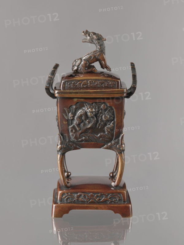 Incense burner kôro with cover, Japanese, Meiji 1868–1912. Incense burner in the shape of a cuboid box on four slightly arched legs standing on a tall rectangular base. Panels with relief decoration of karashishi and peonies. Cover with four holes topped with a qilin. Bronze vessel, 2nd half of the 19th century. Height 22.0 cm; width 9.5 cm; depth 7.5 cm., A small ornate metal vessel in brown and dark bronze tones sits on a pedestal, with a sculpted animal figure mounted on the lid. The vessel has a square body with raised decorative panels showing relief designs, curved handles on either side, and elegantly arched legs that connect to a rectangular base. Surface details include carved floral and scroll motifs and textured patterns, and the overall colors visible are brown, dark bronze, and hints of black.