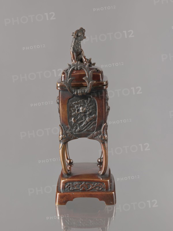 Incense burner koro with cover, Japanese, Meiji 1868–1912. Bronze vessel in the shape of a cuboid box on four slightly arched legs standing on a tall rectangular base. Panels with relief decoration of karashishi and peonies. Cover with four holes topped with a qilin. 2nd half of the 19th century. Height 22.0 cm; width 9.5 cm; depth 7.5 cm., A small ornate object stands against a plain gray background, primarily brown with darker bronze or blackened details; it has a carved animal figure on top in dark bronze tones, decorative curved metalwork in brown and dark bronze beneath it, and a relief panel on the front featuring a floral or botanical motif in dark bronze on a brown base. The piece rests on four curved legs that are brown with darker accents and sit on a rectangular brown platform that has darker carved ornamentation along its front edge. The surface shows polished brown wood tones and darker bronze highlights, and the object casts a faint reflection on the smooth surface below.
