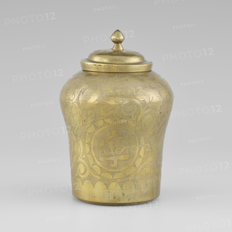 Tin with a lid, tin, early 20th century, Korea, container with widened top, height 15.0 cm, diameter 9.8 cm; ornamented with peonies; Korean art., A single metal container with a lid, rendered in a gold tone, sits against a neutral gray background; the container has a rounded, slightly tapered body with a domed lid topped by a small pointed finial, and its surface is decorated with engraved floral and foliate patterns and textured areas, showing a few darker spots and minor wear on the metal.