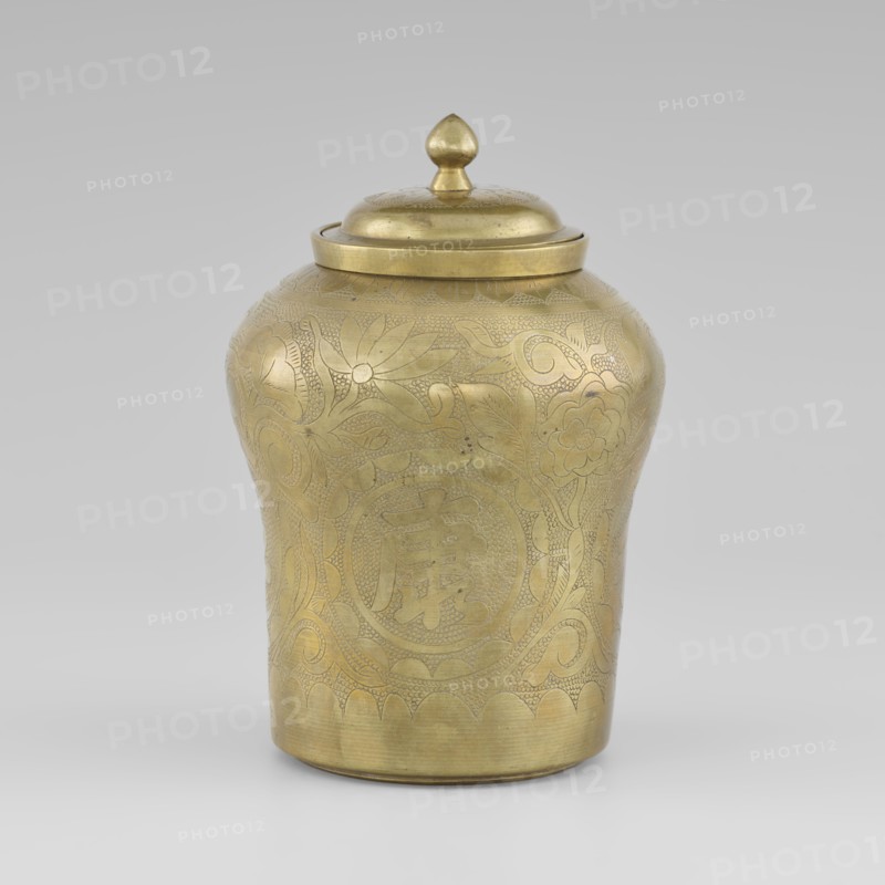 Tin with a lid, tin with the top widened, early 20th century, Korea; container; height 15.0 cm, diameter 9.8 cm; decorated with peonies; Korean art; plant ornament;, A single lidded metal container with a rounded body and slightly flared base sits against a neutral gray background; the container is golden in color with a polished sheen and features etched decorative patterns of flowers, leaves, and swirling motifs covering its surface; the lid is domed with a teardrop-shaped finial on top, also golden, and the decoration includes areas of fine stippling and engraved line work; the overall color palette is gold with subtle variations in tone where light and shadow create highlights and darker areas.