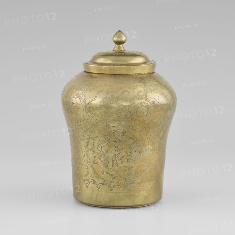 Tin with a lid; tin with the top widened; container, height 15.0 cm, diameter 9.8 cm; early 20th century, Korea; Korean art; Korean culture; ornament: plant peonies;, A brass jar with a lid, showing an overall golden color with engraved decorative patterns covering its rounded body and lid; the jar has a bulbous upper section tapering to a narrower base, a domed lid topped by a small finial, and visible engraved motifs of flowing curved lines and stylized leaf and floral shapes; the background is a neutral light gray.
