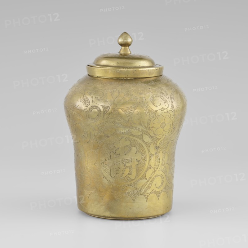 Tin with a lid; tin; early 20th century; Korean; container with widened top; height 15.0 cm; diameter 9.8 cm; decorated with peony plant ornament., A brass lidded jar with a rounded body and a domed lid topped by a teardrop-shaped finial fills the frame, showing an overall golden color with a slightly aged patina. The surface of the jar is decorated with incised floral and foliate patterns, including stylized flower motifs and curling vine-like designs, all rendered in the same golden tone with subtle variations in brightness where the metal is more polished or shaded. The jar has a gently scalloped border near the base and a smooth rim beneath the lid. The background is a neutral light gray, and the jar sits on a similarly light surface.