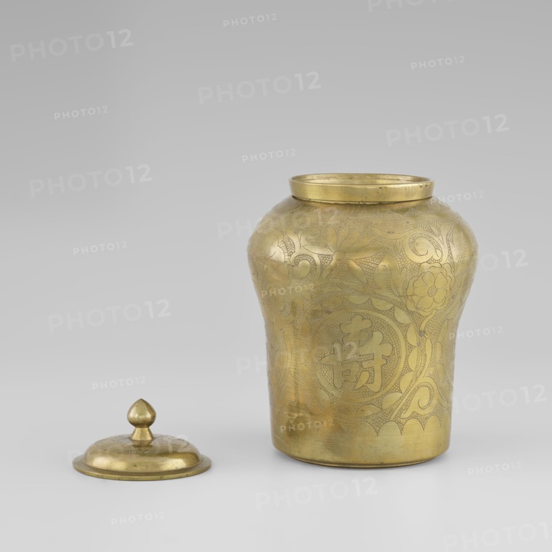 Tin with a lid; tin; plant ornament with peonies; Korean; early 20th century; container; height 15.0 cm, diameter 9.8 cm., A brass-colored metal jar with a rounded body and flared rim sits on a light neutral surface, accompanied by its separate lid placed nearby. The jar's exterior shows engraved floral and swirling decorative patterns covering much of its surface, with varied textures from smooth reflective areas to finely incised detailing. The lid is dome-shaped with a smooth knob finial and has a matching brass color and subtle engraved decoration. The background is a gradient of light gray tones. The visible colors are brass/golden, light gray, and subtle darker gray shadows.
