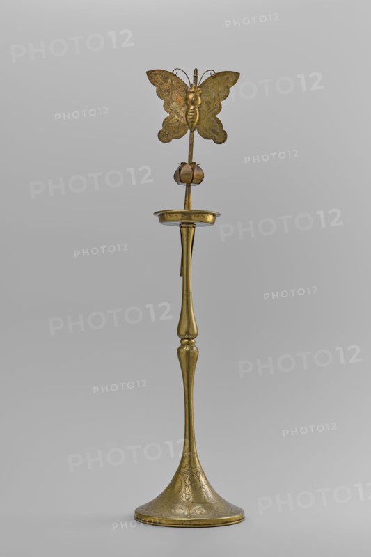 Candlestick, early 20th century, Korea; candlestick on a round base transitioning into a two-baluster shaft terminating in a flat, round sleeve with a smaller one in the shape of a multi-petaled flower bud placed in the center; decorated with butterflies and flowers; from a pair other no. SKAZsz 2509; height 39.7 cm., A tall, slender metallic object with a warm golden color stands against a plain light gray background; its flared base is decorated with etched swirling patterns, the narrow stem rising with smooth, rounded contours to a shallow dish-like platform, above which a thin rod supports a stylized flower bud and, at the top, a symmetrical butterfly-shaped ornament with delicate etched wing patterns and curled antennae, all sharing the same golden hue with subtle variations in sheen and small darker areas from patina.