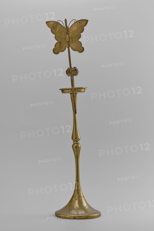 Candlestick, early 20th century, Korea; candlestick on a round base transitioning into a two-baluster shaft terminating in a flat, round sleeve with a smaller one in the shape of a multi-petaled flower bud placed in the center; decorated with butterflies and flowers; from a pair; height 39.7 cm., A tall metallic object with a slender, elongated stem and a flared, rounded base etched with decorative patterns, topped by a small shallow dish and a vertical rod that culminates in a butterfly-shaped ornament; the surface appears predominantly brass-colored with areas of darker patina and subtle variations in tone, set against a neutral light-gray background.