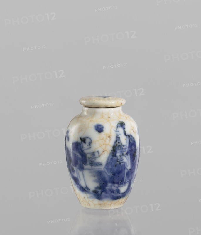 Flask - snuffbox with stopper, maker, repossession noted. Ceramics, Chinese, figural scenes, blue-and-white porcelain, cracklework decoration. The flask is short and wide with a short neck and cracklework embedded in yellow paint. The porcelain stopper is flat. The glaze is bluish, transparent, with minor impurities. Decoration is underglaze painted with cobalt. The stopper and flask are decorated with cracklework. 19th century, China; vessel; height 6 cm, diameter 4 cm., A small ceramic jar with a rounded body and a fitted lid sits against a neutral gray background; the jar is predominantly white with visible age-related hairline crack patterns in a tan color and features a painted blue scene wrapping around its body showing human figures and abstract shapes; the lid is similarly white with tan cracking and a small dark spot on top, and the base of the jar shows a subtle reflection on the surface beneath it.
