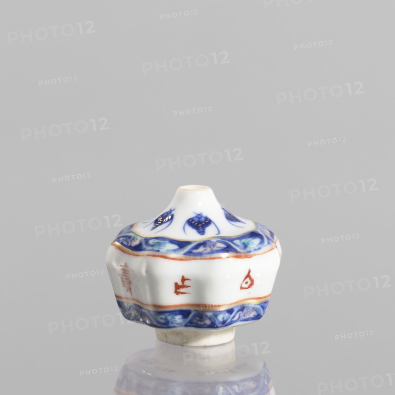 Spice vessel, ceramics, blue and white porcelain, 19th century, China; subject: bats; function: spice vessel No stopper; dimensions: height 4.65 cm, diameter 5.5 cm., A small white ceramic object with a rounded, flared shape sits against a light gray background, featuring decorative bands of blue and orange around its midsection and base, and blue floral-like motifs on the upper section; the piece has smooth glossy surfaces, a short central opening at the top, and a slightly irregular foot with a beige underside, and a reflection is visible beneath it.