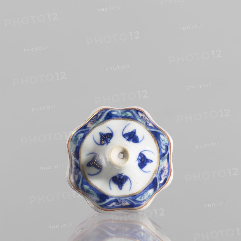 Spice vessel, 19th century China, blue-and-white porcelain; vessel no stopper; height 4.65 cm, diameter 5.5 cm; decorated with bats; used for spices., A small scalloped ceramic object with a glossy white base and a central raised hole, decorated with a blue border featuring floral-like motifs and several blue stylized shapes on the white surface, with touches of gold accenting parts of the design and a thin reddish-brown rim, all reflected faintly on the glossy surface beneath; visible colors include white, various blues, gold, reddish-brown, and a hint of green.