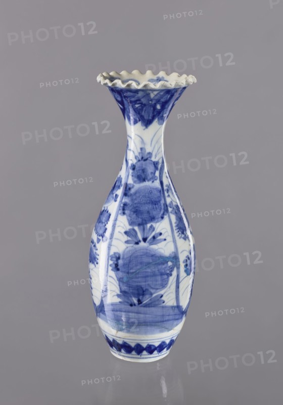 Vase, ceramics, blue and white porcelain, Japanese, 19th/20th century Japan, plant ornaments, vessel, height 36.6 cm, diameter 14 cm, A ceramic vase stands against a plain gray background; the vase has a narrow neck flaring into a ruffled rim and a bulbous body tapering to a small base, decorated with blue painted floral and abstract motifs on a white ground, showing brushstroke textures and areas of darker and lighter blue, with the rim edge appearing off-white and the overall color palette including blue, white, gray, and a touch of beige.
