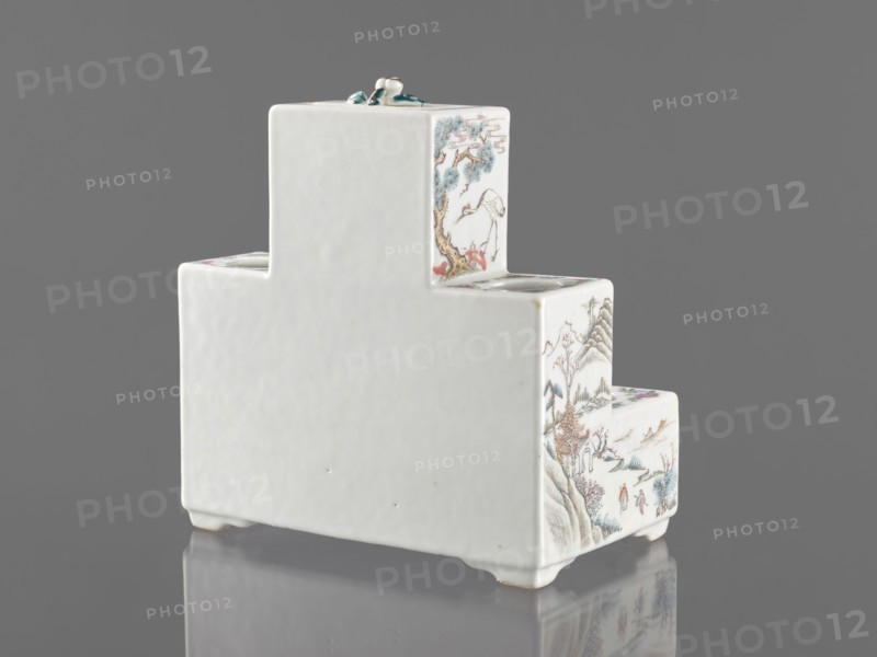 Pedestal for a writing set three-stage, ceramics with colored porcelain in Qing style late 19th century, China. Decorated with flowers, figural scenes, butterflies, and landscape motifs; raised central part. Function: writing accessories. Dimensions: height 18.9 cm, width 17.9 cm, depth 11.0 cm., A white stepped ceramic object with a glossy finish sits on a reflective surface against a gray background; the visible faces feature delicate painted scenes in muted colors including blue, green, brown, pink, and red, showing natural elements like trees, mountains, and birds, and small human figures in traditional clothing; the top has a small sculpted decorative element in green and white, and the object rests on short feet with slight shadows and reflections beneath.