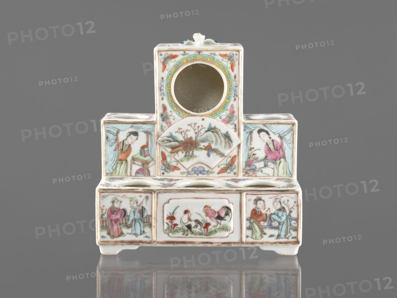 Pedestal for a writing set three-stage, colored porcelain, late 19th century, China; Qing style Qing, 1644–1911. Decorated with ceramics flowers, butterflies, landscape motifs and figural scenes; raised central part. Function: writing accessories. Dimensions: height 18.9 cm; width 17.9 cm; depth 11.0 cm., A small ceramic object with a stepped rectangular form sits against a gray background, predominantly white with painted decoration in colors including pink, green, blue, yellow, orange, brown, and black. The piece has a central circular opening near the top and a few round openings along the midline, and is supported by short feet. Painted panels on the front and sides depict several human figures wearing flowing robes in pink, green, blue, and red tones, scenes of people interacting, and a pair of roosters amid grass and flowers. Other panels show a landscape with trees and hills and decorative floral and butterfly motifs in pastel shades. The painted outlines and details are rendere...