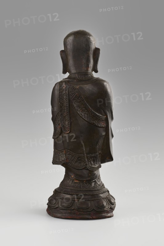 Monk, Papal, Chinese art, Chinese culture, Qing 1644-1911, figurine, Buddhist art depicting a monk/arhat Luohan, lotus throne with round low lotus pedestal and lotuses, 19th/20th century, China, sculpture, height 24.7 cm, diameter 9 cm., A dark brown carved figure seen from behind standing on a decorative rounded base with layered petal-like shapes, the figure wearing a draped garment with patterned borders and engraved decorative details, a smooth rounded head with visible ears, and small areas of reddish-brown showing through the worn surface; the background transitions from light gray at the bottom to a mid-gray at the top.