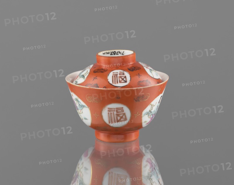 Bowl with a lid, Qing dynasty Tongzhi period, 1862–1874, Chinese, colored porcelain, ceramics; decorated with bats and swastikas and genre scenes, inscription/decoration note: In the background of the decoration loosely placed bats and swastikas. Vessel; height 8.6 cm, diameter 10.3 cm., A small lidded ceramic bowl with a rounded foot and flared rim sits centered on a reflective surface, showing a mirrored image below. The bowl is primarily a warm orange color with white circular panels around its body and lid that contain decorative motifs. There are accents of gold outlining small cloud-like shapes and other tiny decorative marks on the orange background. Within the white panels are painted scenes and figures in colors including green, pink, purple, and pale blue, with some black or dark brown fine lines for detailing. The interior visible at the rim is white, and the overall background behind the object is a flat gray.