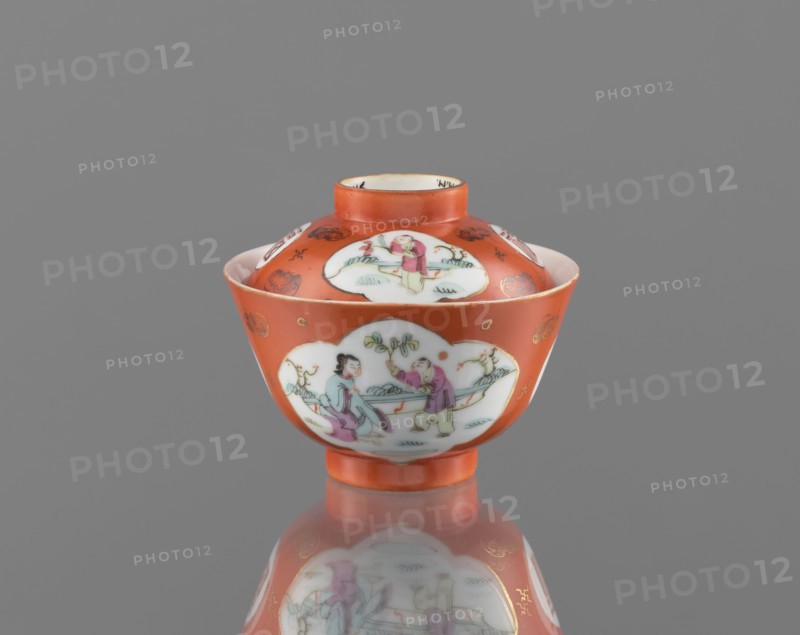 Bowl with a lid;, ; Qing dynasty 1644–1911, Tongzhi period 1862–1874; Chinese; colored porcelain ceramics; vessel decorated with bats and swastikas and genre scenes, with bats and swastikas loosely placed in the background of the decoration; height 8.6 cm, diameter 10.3 cm., A small lidded bowl with a rounded body and short foot, painted predominantly in orange with white cartouche-shaped panels showing scenes of human figures and foliage; the lid and body feature several of these white panels bordered by orange, with delicate painted details in pink, green, blue, and black, and small gold decorative accents on the orange background; the interior rim of the lid appears white and the bowl sits on a reflective grey surface that mirrors its colors and shapes, set against a plain grey backdrop.