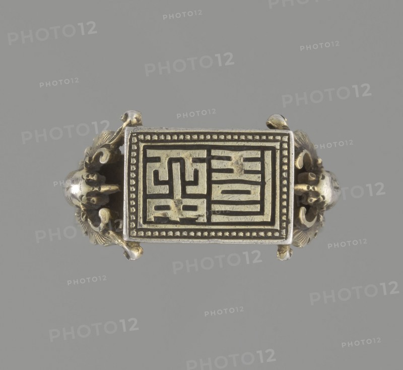 Ring, Chinese, Qing 1644–1911, 19th century. Jewelry; hand ornament. Shield with ideographic signs supported by two bats motif; bats iconography. Diameter 2 cm. ., A rectangular metallic object with ornate curved sides rests against a plain gray background; the rectangle has a raised border with a dotted inner edge and an inset panel featuring a pattern of raised linear shapes, and the curved side elements include decorative scroll-like and floral motifs with rounded bulbous forms. The visible colors are metallic gold, silver, and gray.