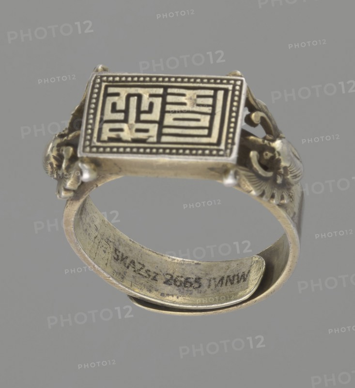 Ring, Chinese, Chinese art, Qing 1644–1911, body ornament/jewelry/hand ornament, motif: shield with ideographic signs supported by two bats, 19th century, China, diameter 2 cm, A metal ring photographed against a plain gray background, showing a rectangular top with a raised patterned design and a dotted border, ornate sculpted sides with decorative motifs, and a curved band with visible surface wear; the visible colors are silver, gray, and muted gold.