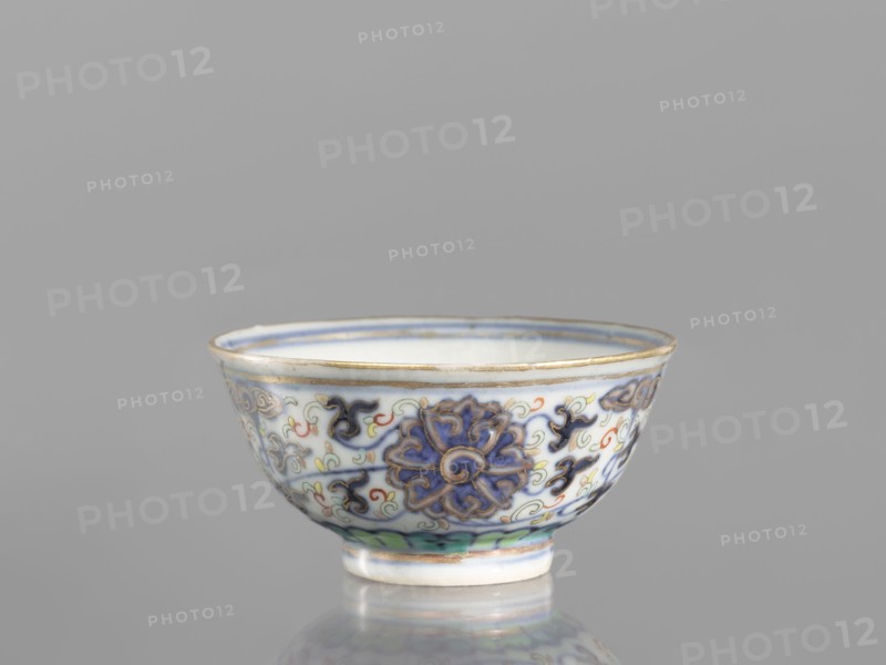 Bowl, Chinese, Qing dynasty 1644–1911, 19th century; colored porcelain vessel with plant and floral ornaments including lotuses and lotus tendril motif, floral motifs on the reverse of the rim; diameter 6.8 cm., A small ceramic bowl sits on a reflective surface against a neutral gray background, showing a rounded rim and a short foot; the bowl is predominantly white with decorative motifs in blue and purple, accented with green, yellow, red, and black, and the rim and foot edges have a thin gold or brass-colored band; the exterior displays a repeating floral or scroll-like pattern in deep blue and purple with sinuous outlines and small accent swirls in red and yellow, and there is a green band near the foot.