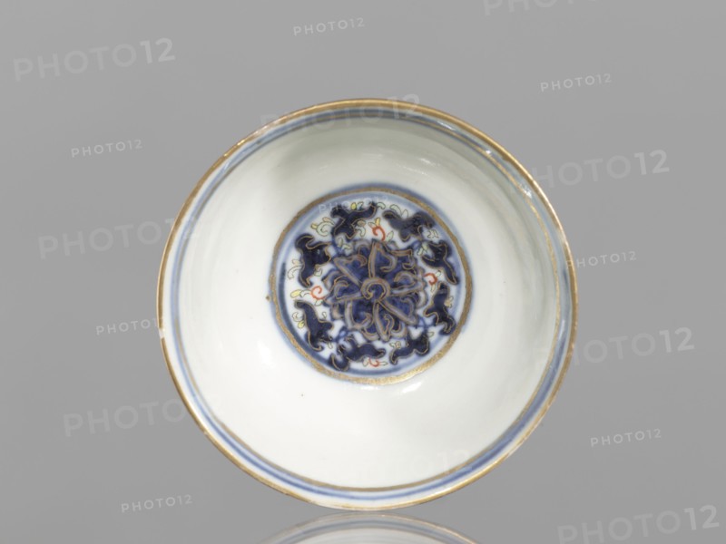 Bowl, 19th century, China; Qing dynasty 1644–1911; colored porcelain vessel, diameter 6.8 cm; decorated with plant and floral ornaments—flowers, lotuses, and lotus tendril motif—with floral motifs on the reverse of the rim., A shallow round bowl viewed from above with a glossy white interior and a decorative circular medallion at the center featuring a floral-like pattern in dark blue with small accents of red and pale yellow, surrounded by concentric bands in blue and gold; the rim has a thin gold edge and a thin blue line just inside it, and the background is a uniform light gray.