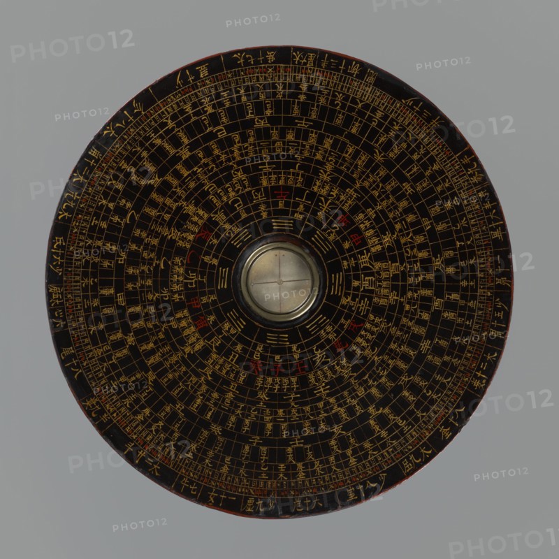 Feng shui compass luopan, Chinese, Qing 1644–1911, 19th century. San Yuan Luo Pan also known as yi pan or jiang pan containing the ring for the 64 hexagrams of the I Ching and a 24-direction ring; used by practitioners of the Three Cycles System of Feng Shui including Flying Stars Time Space. Materials/format: navigational/astrological measuring instrument compass, diameter 17.0 cm. Subjects/themes: astrology, calendars, Chinese Zodiac., A round, flat object viewed from above with concentric circular patterns and a central raised circular element resembling a glass-covered disk; the main surface is dark brown to black with a complex arrangement of thin lines and small markings in gold and occasional small areas of red, the outer rim showing a slightly worn edge with hints of reddish-brown, and the background is a uniform light gray.
