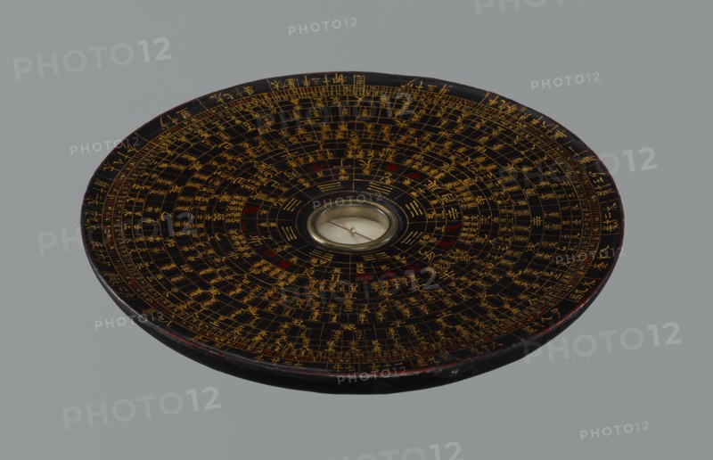 Feng shui compass luopan, Chinese, Qing 1644–1911, 19th century. San yuan luo pan also known as yi pan or jiang pan with the ring for the 64 hexagrams of the I-Ching and a 24-direction ring; used by practitioners of the Three Cycles System of Feng Shui including Flying Stars Time Space. Function: navigational/astrological measuring instrument. Diameter 17.0 cm., A circular, flat object with concentric rings of fine markings and grid-like divisions across its dark surface, featuring a central round glass-covered element with a small needle; the surface shows a pattern of primarily gold lines and symbols with occasional red accents on a black background, and the rim has a worn dark edge with hints of brown; the overall background is a uniform gray.