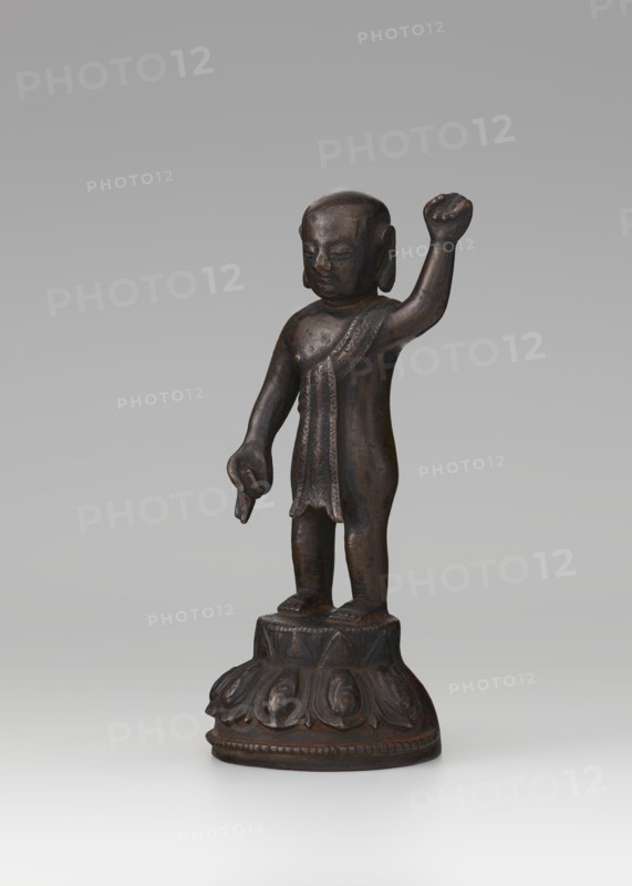 Young Buddha, unknown sculptor, repossession Krakow, Buddha iconography, figurine on lotus throne, Chinese art, Chinese Buddhism, children, Qing 1644–1911, Buddhist art, 19th/20th century, China, sculpture, height 16.7 cm, A small dark brown bronze statue of a standing human figure on a round ornamental base, the figure with a smooth rounded head and simplified facial features, one arm raised with a closed hand and the other arm lowered with fingers extended, wearing a draped garment over one shoulder with textured decorative details, the legs close together and feet positioned on a tiered platform that has petal-like reliefs; background is a gradient of light gray.