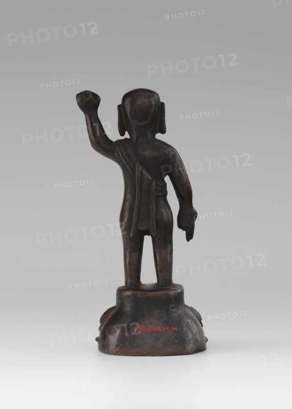 Young Buddha, unknown sculptor, gift, repossession Krakow, Buddha iconography, figurine on lotus throne, Chinese art, Chinese culture, Chinese Buddhism, children, Qing 1644–1911, Buddhist art, 19th/20th century, China, sculpture, height 16.7 cm, A small dark brown statue viewed from behind standing on a rounded base, with a raised arm and a lowered arm, a draped cloth over one shoulder and down the back, short legs and rounded head, and visible colors including dark brown, hints of black, and a neutral gray background.