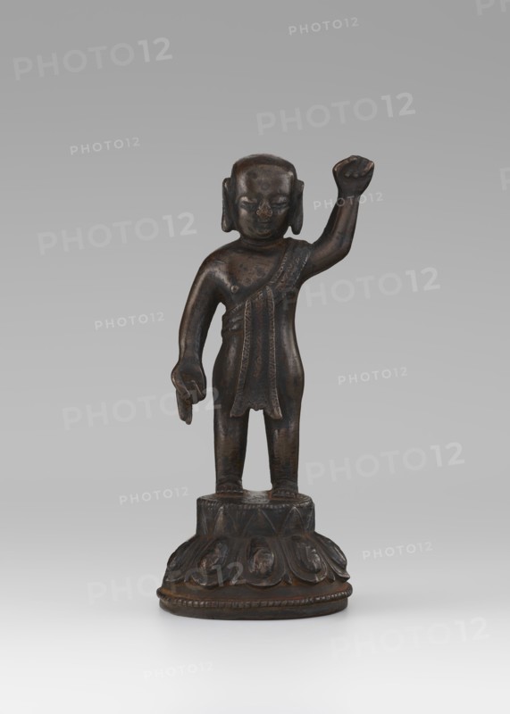 Young Buddha, unknown sculptor, gift, repossession Krakow, Buddha iconography, figurines, lotus throne iconography, Chinese art, Chinese culture, Chinese Buddhism, children, Qing 1644–1911, Buddhist art, 19th/20th century, China, sculpture, height 16.7 cm, A small dark brown metal statue of a standing figure on a round decorated base is visible; the figure has a raised right arm and a lowered left arm with a hand gesture, a short garment draped over one shoulder, stylized hair or head covering, and a smooth patina showing darker and lighter brown tones, set against a neutral gray background.